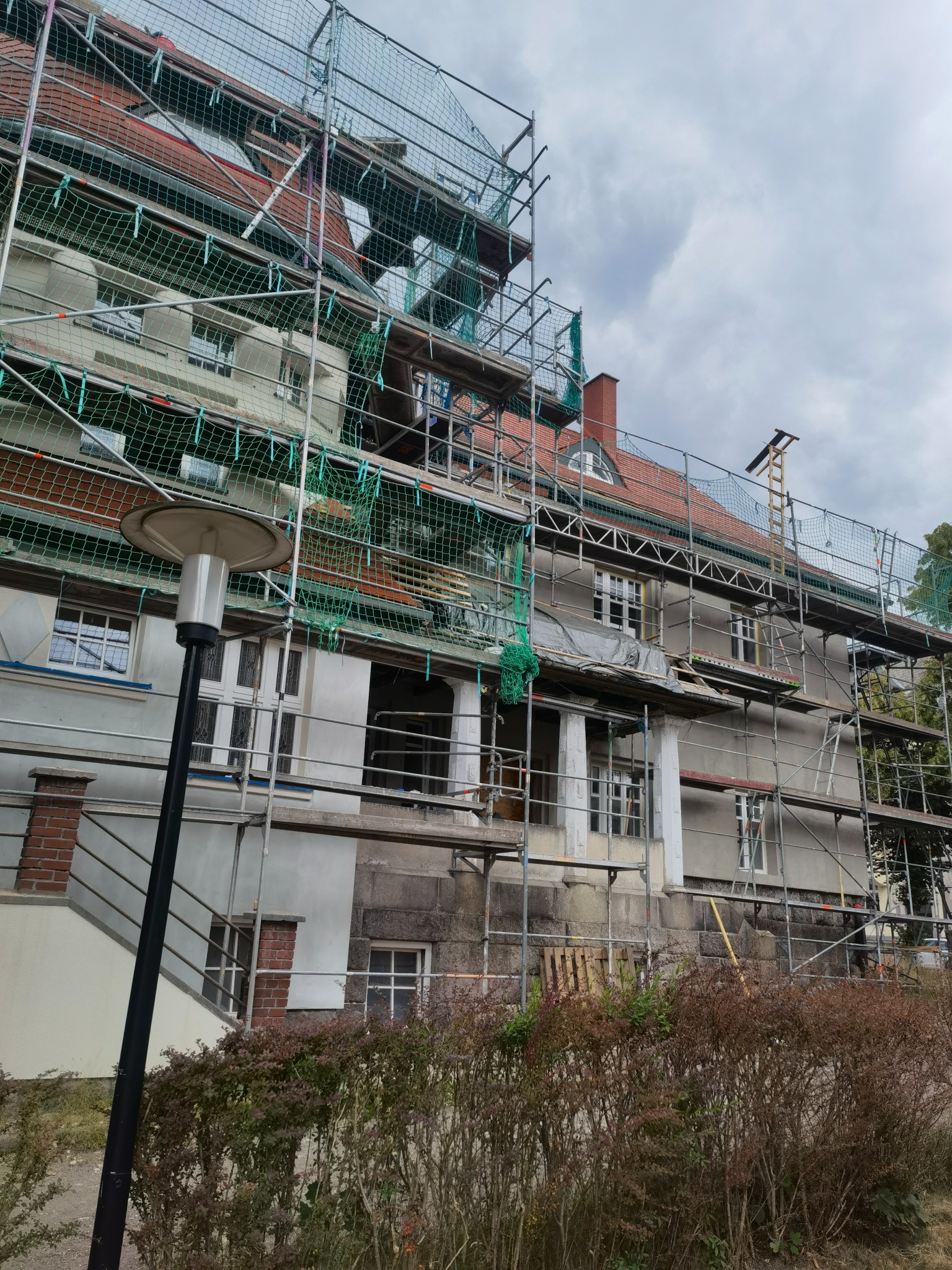 Budynek w trakcie prac elewacyjnych, otoczony rusztowaniami i siatką ochronną, widoczna lampa uliczna w pierwszym planie, zachmurzone niebo.