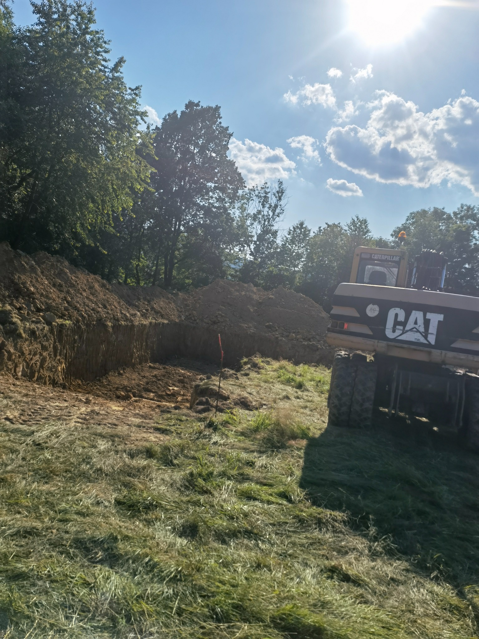 Żółta koparka CAT częściowo widoczna obok wykopu w ziemi, trawa na pierwszym planie, drzewa w tle, słoneczny dzień.