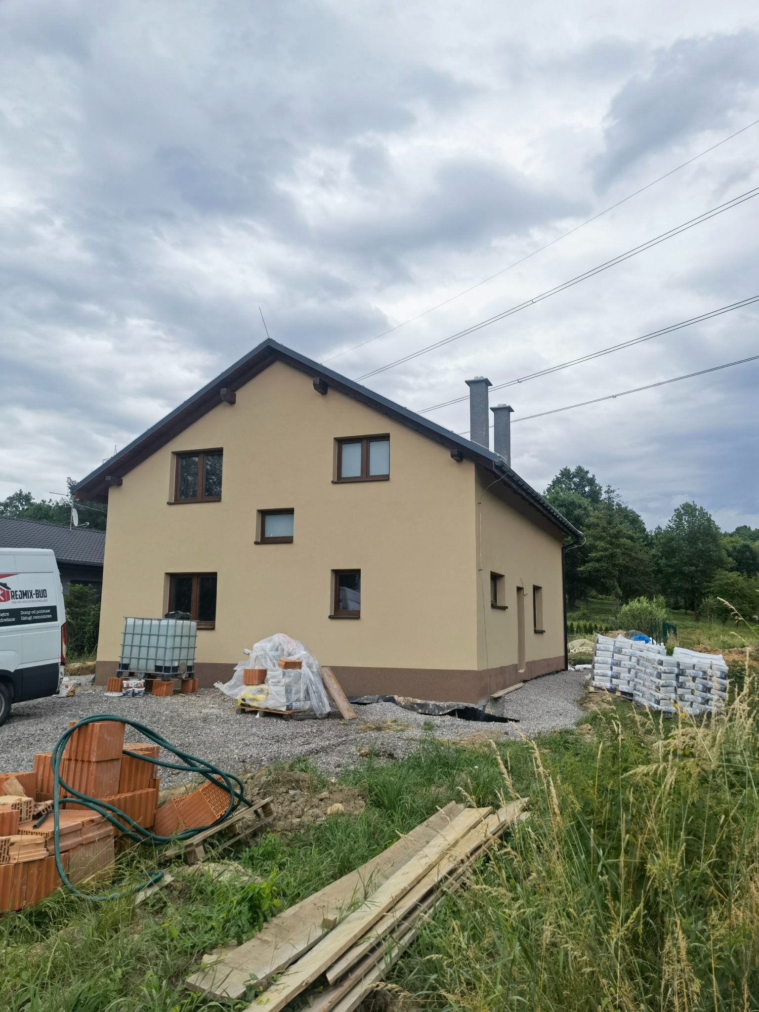 Dwukondygnacyjny dom z jasnobeżową elewacją, ciemnobrązowym pasem u podstawy i ciemnymi oknami, częściowo otoczony materiałami budowlanymi i wysoką trawą, pod pochmurnym niebem.