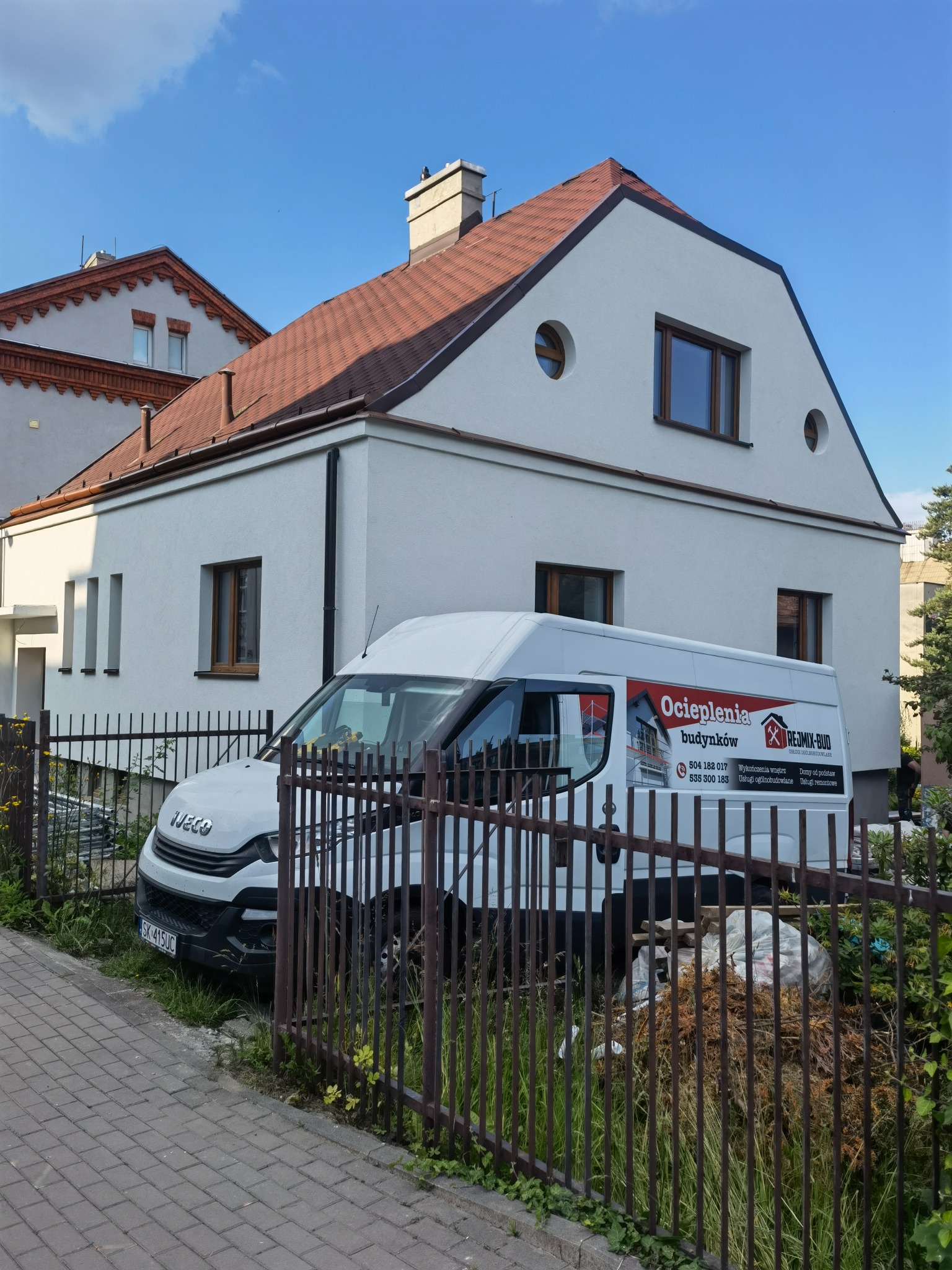 Biały dom jednorodzinny z nową elewacją, brązowym dachem i oknami, częściowo zasłonięty przez biały samochód dostawczy z reklamą firmy budowlanej, widok zza metalowego ogrodzenia.