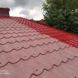 Czerwony dach z blachy trapezowej w trakcie mycia, z widocznym kominem zabezpieczonym taśmą i częściowo umytą powierzchnią, na tle zieleni i pochmurnego nieba.