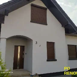 Biały dom jednorodzinny z brązowymi roletami zewnętrznymi i ciemnym dachem po renowacji, widoczne numer domu '3' i fragment zielonego krzewu.