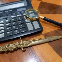Kalkulator Citizen SDC-875A, lupa z pozłacaną ramką i ozdobny nożyk do papieru z motywem liści dębu leżą na drewnianym biurku w szachownicę.