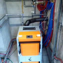 Nowoczesny piec CO z pomarańczowymi drzwiczkami w kotłowni, widoczne rury instalacji grzewczej w kolorach czerwonym i niebieskim, oraz szare rury wentylacyjne pod sufitem.