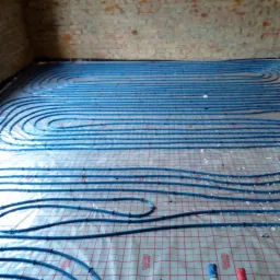 Niebieskie rury ogrzewania podłogowego ułożone spiralnie na folii z czerwoną siatką, widok z góry, surowa ceglana ściana w tle.