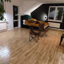 Pok&oacute;j z drewnopodobną podłogą, zielonym ł&oacute;żkiem z pikowanym zagł&oacute;wkiem i czarną ścianą, oświetlony lampą ścienną, z dwoma krzesłami turystycznymi na pierwszym planie i roślinami doniczkowymi.