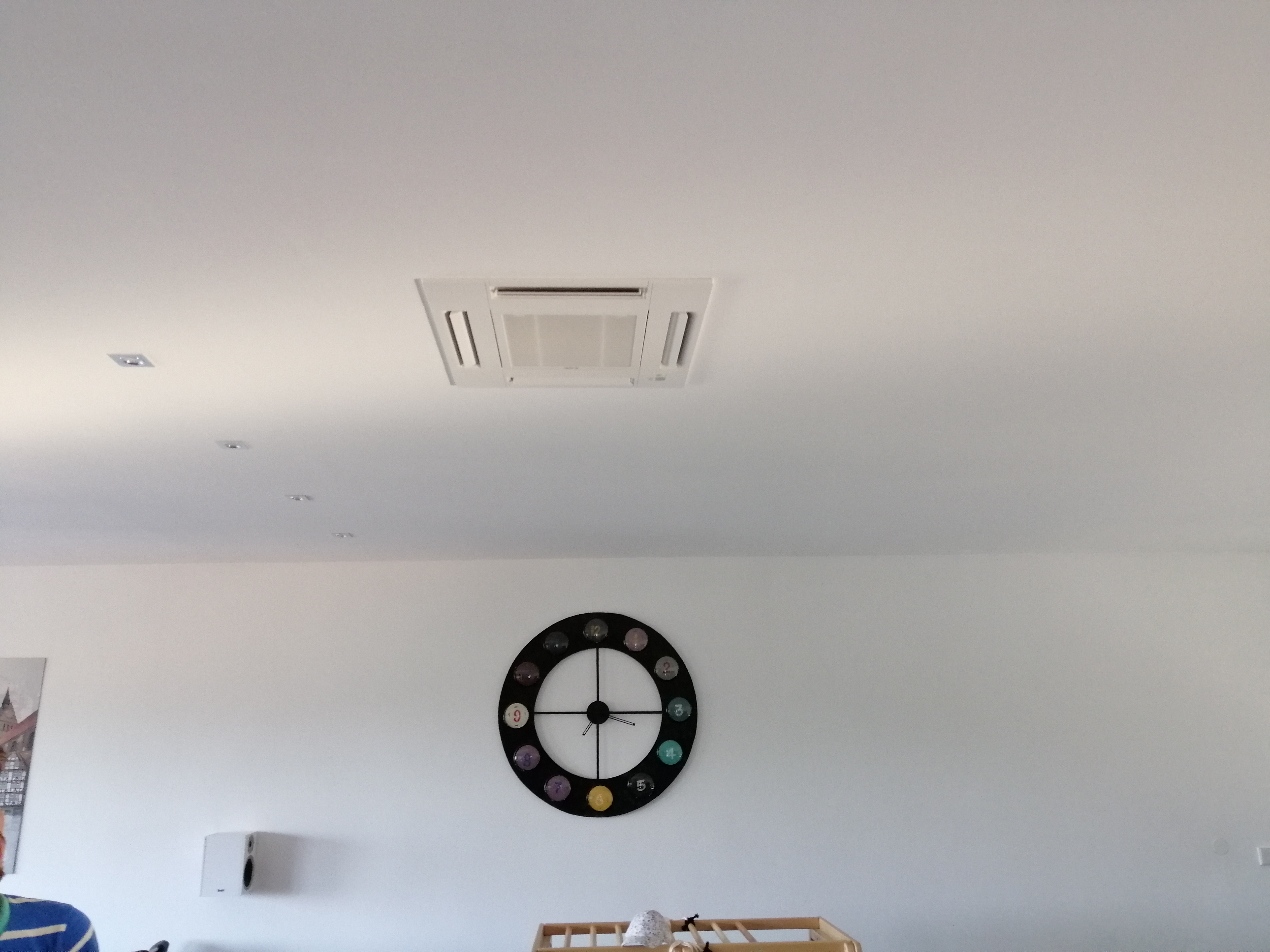 Kasetonowa jednostka klimatyzacji zamontowana w suficie podwieszanym w jasnym pomieszczeniu mieszkalnym z zegarem ściennym i elementami wyposażenia wnętrza.