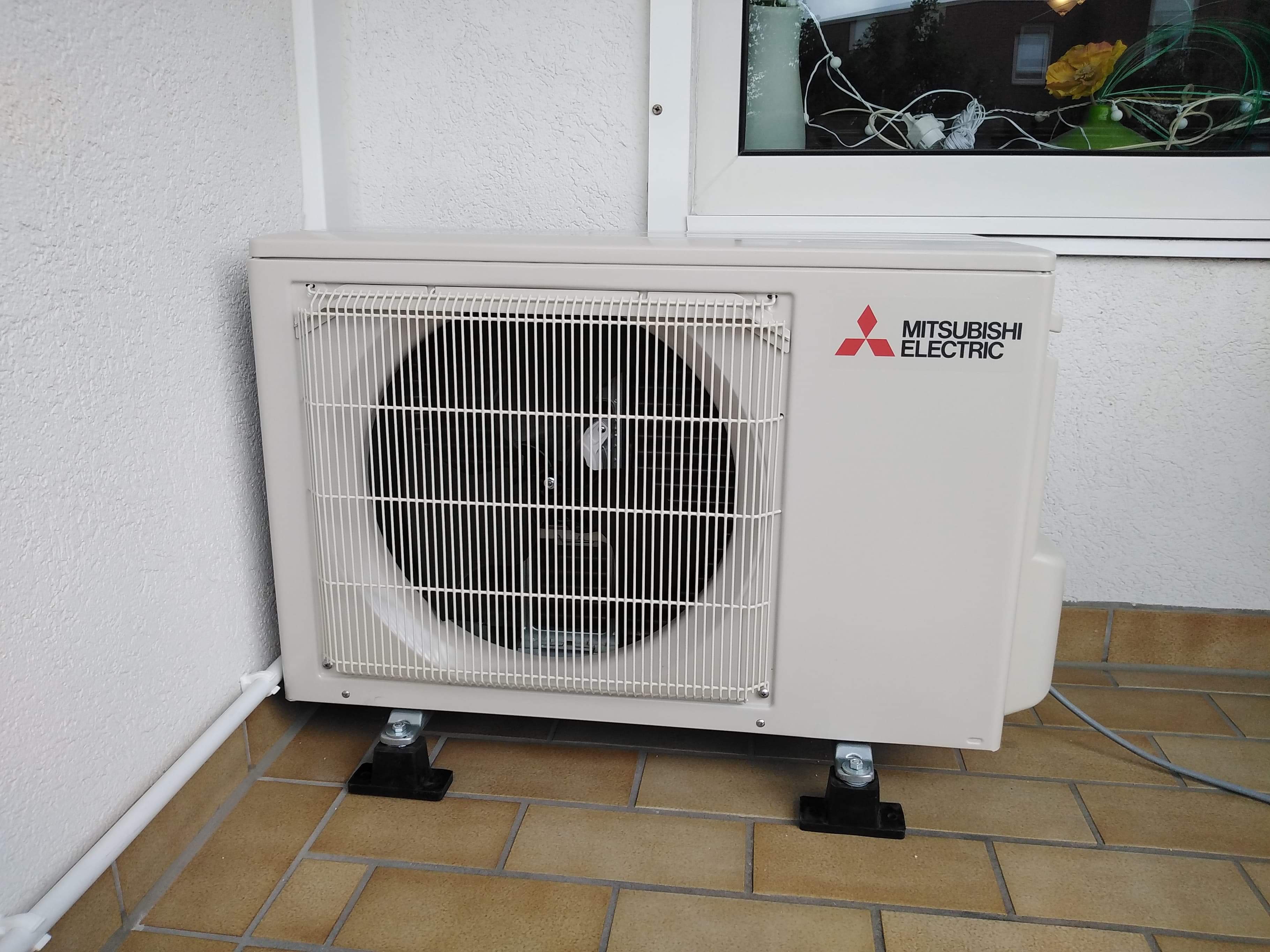 Zewnętrzna jednostka klimatyzacji Mitsubishi Electric zamontowana na czarnych wspornikach na brązowej podłodze z płytek, widok z przodu, okno z dekoracją w tle.
