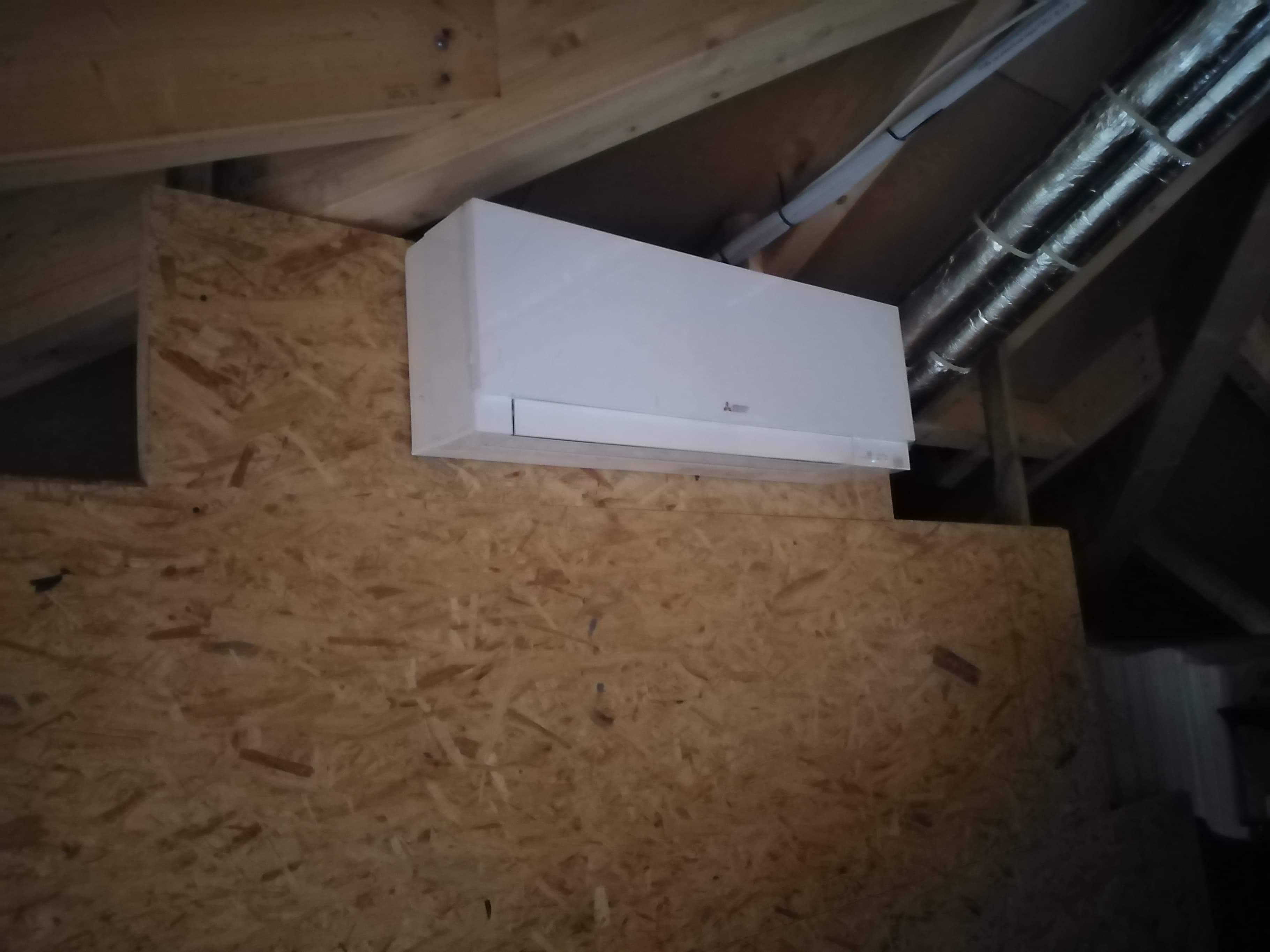 Biała jednostka wewnętrzna klimatyzacji marki Mitsubishi zamontowana na ścianie z płyty OSB na poddaszu, widoczne rury instalacyjne pod sufitem.