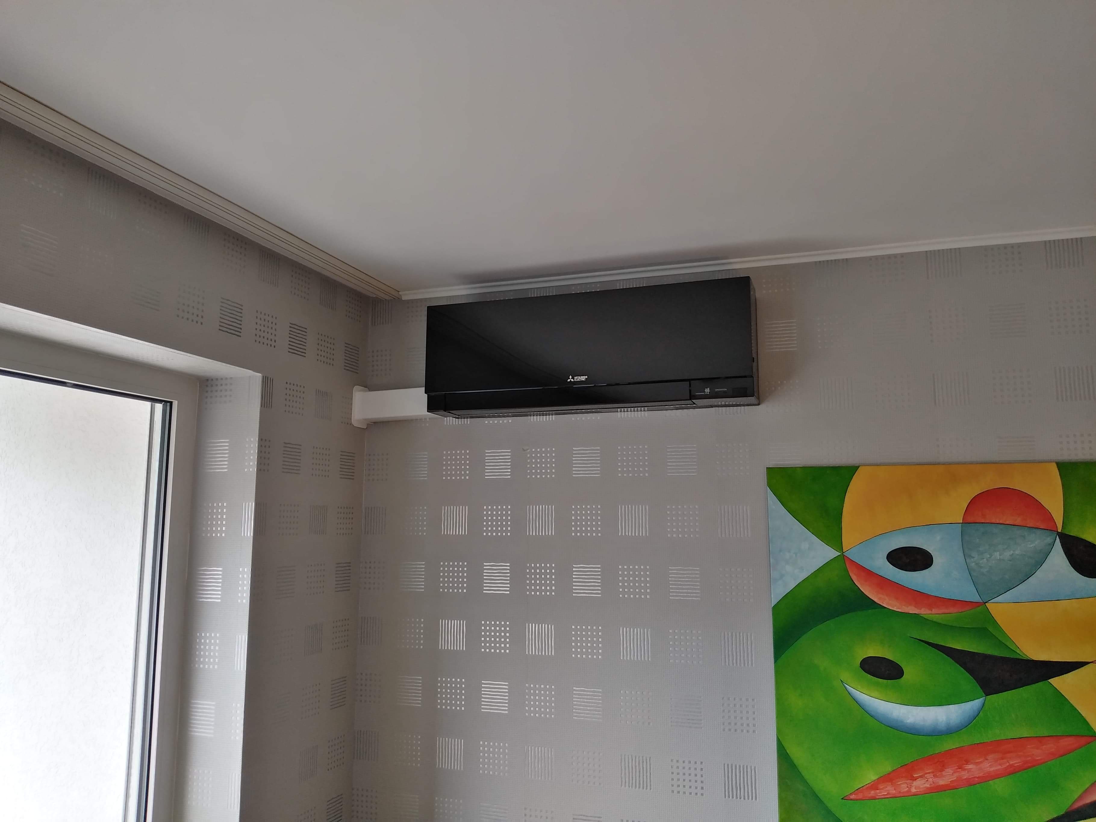 Czarny klimatyzator Mitsubishi zamontowany na ścianie w pokoju z tapetą w geometryczne wzory, obok okna i abstrakcyjnego obrazu.