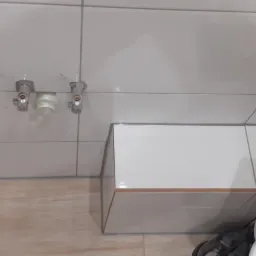 Częściowo wykończona łazienka z białymi płytkami, widoczne zawory wodne i fragment toalety.