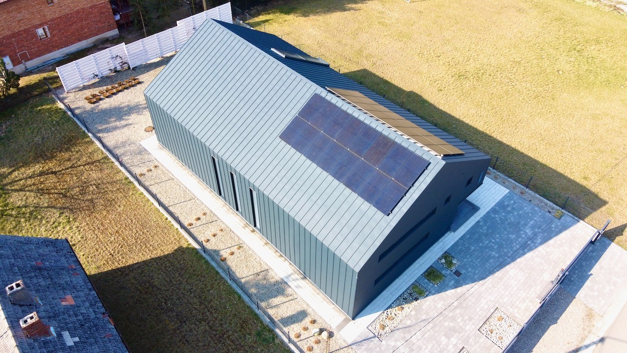 Widok z góry na nowoczesny dom z szarą blachą na dachu i panelami słonecznymi; obok domu utwardzony podjazd i trawnik.