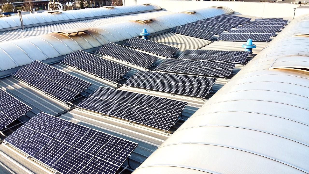 Rząd paneli słonecznych na falistym dachu przemysłowym z niebieskimi odpowietrznikami, widok z góry.