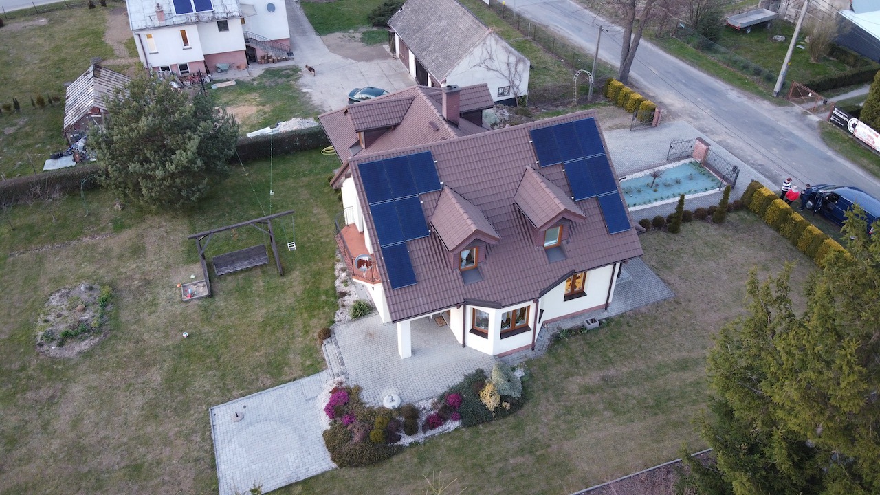Widok z drona na dom jednorodzinny z brązowym dachem i panelami słonecznymi, otoczony ogrodem z huśtawką i ozdobnymi krzewami.
