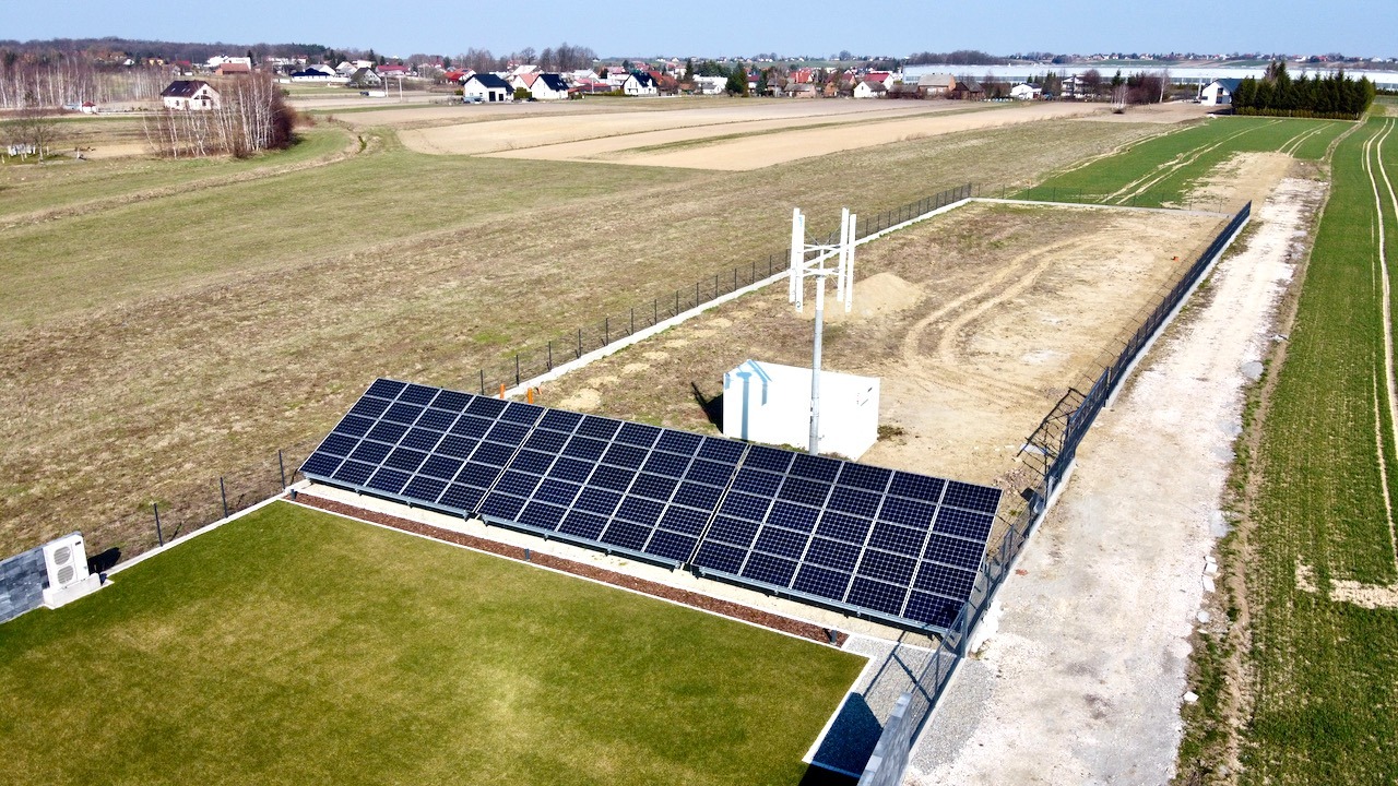 Widok z góry na panele słoneczne zamontowane obok trawnika i wiatraka na tle wiejskiego krajobrazu.