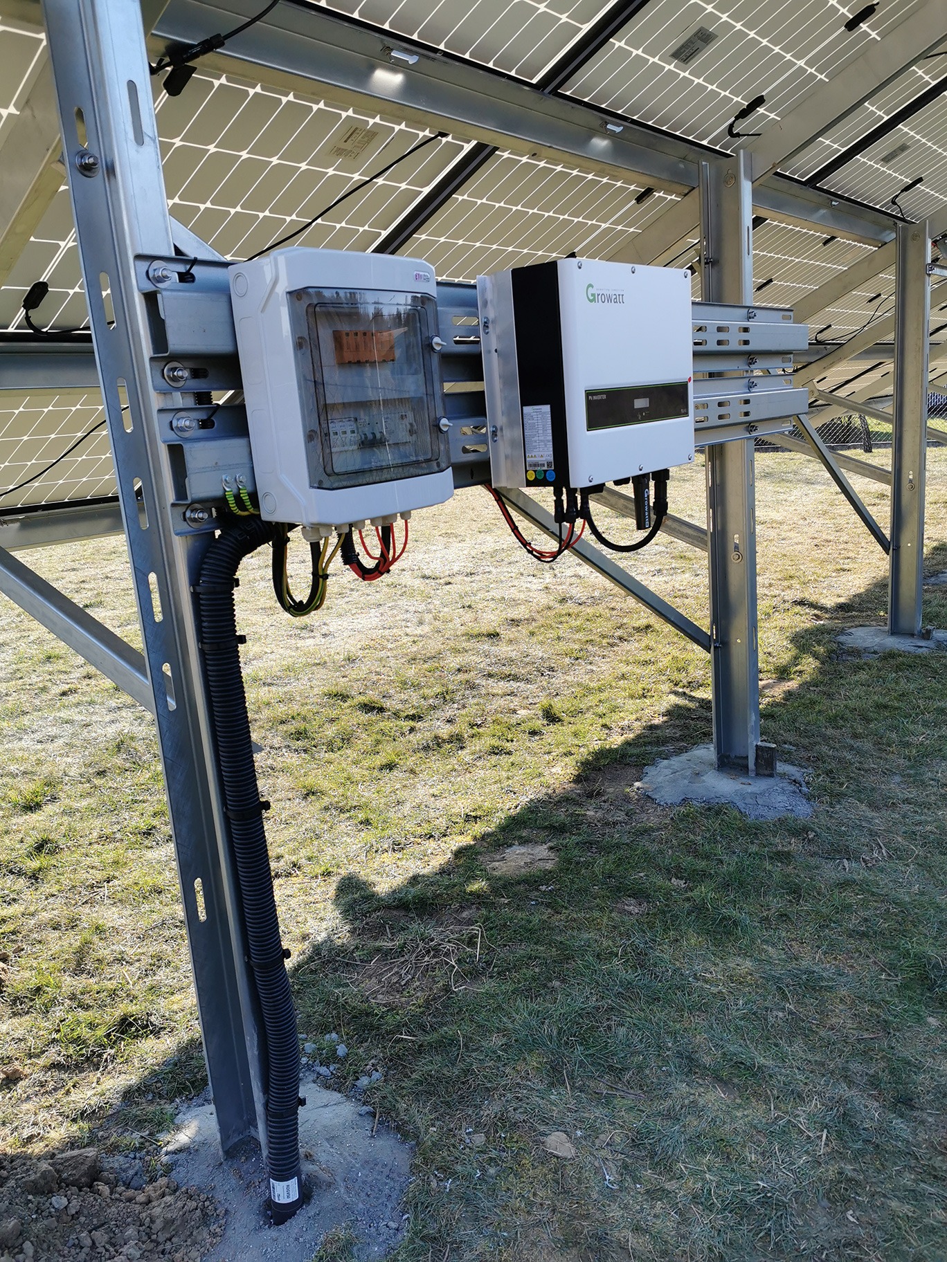 Instalacja paneli słonecznych z widoczną skrzynką rozdzielczą i inwerterem Growatt, zamontowanymi na metalowej konstrukcji na trawiastym terenie.