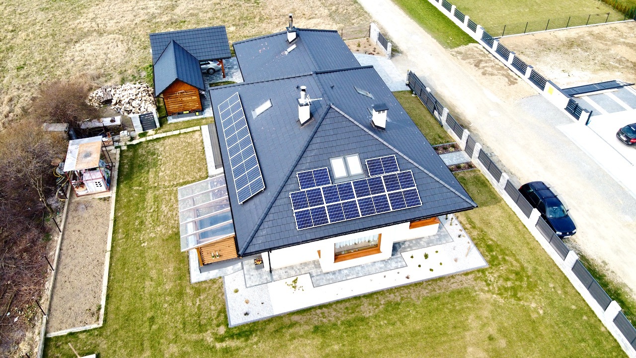 Widok z drona na dom jednorodzinny z panelami słonecznymi na dachu i wbudowaną szklarnią, otoczony zielonym trawnikiem i ogrodzeniem.