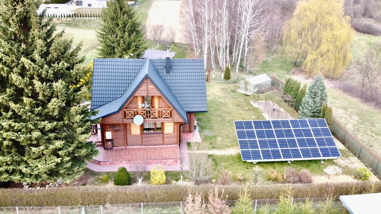 Drewniany dom z ciemną dachówką i panelami słonecznymi w ogrodzie, widok z góry.