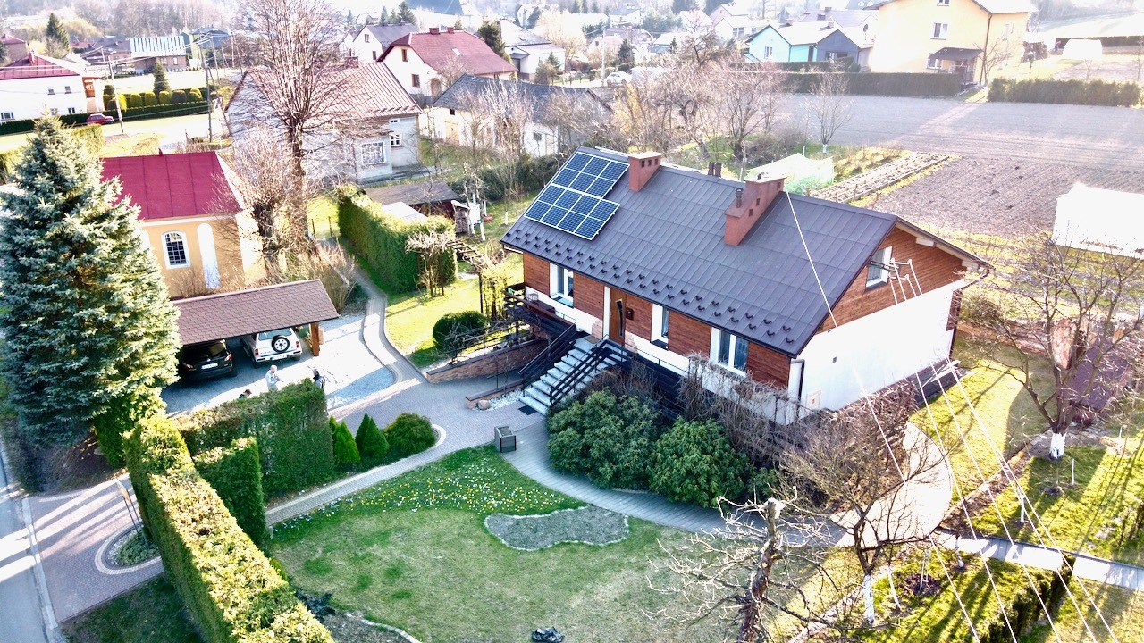 Dom jednorodzinny z brązową dachówką i panelami fotowoltaicznymi na dachu, widok z góry na zadbany ogród i okoliczne budynki w słoneczny dzień.