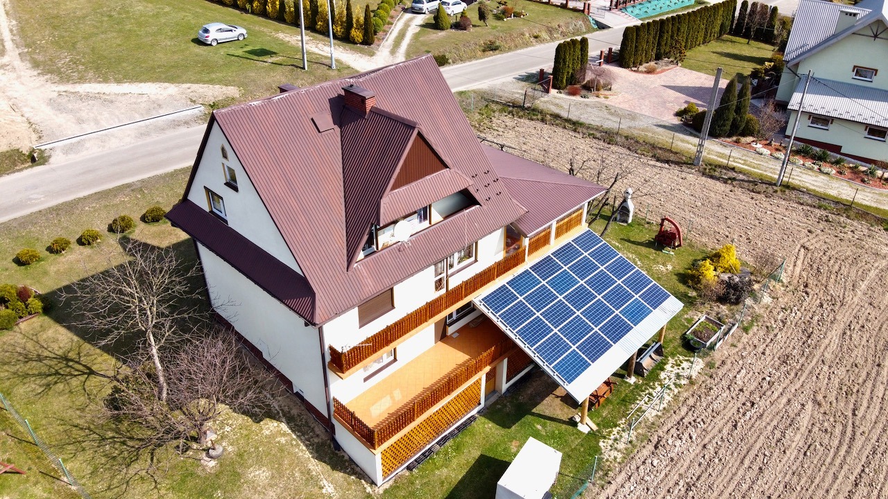 Widok z góry na dom jednorodzinny z brązowym dachem i panelami fotowoltaicznymi zamontowanymi na zadaszeniu tarasu.