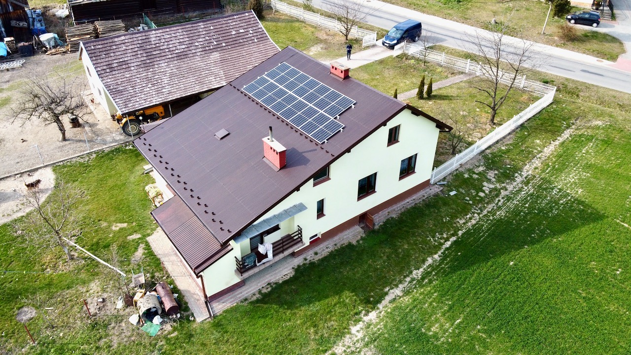 Widok z góry na dom jednorodzinny z brązowym dachem, na którym zamontowane są panele słoneczne; w tle widoczny budynek gospodarczy i ulica.