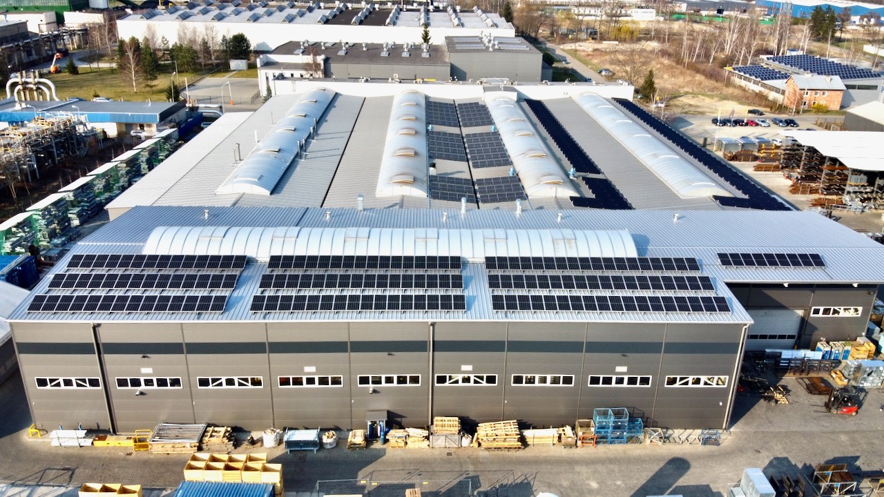Widok z góry na duży, szary budynek przemysłowy z zainstalowanymi panelami słonecznymi na dachu, otoczony innymi budynkami i placem składowym.