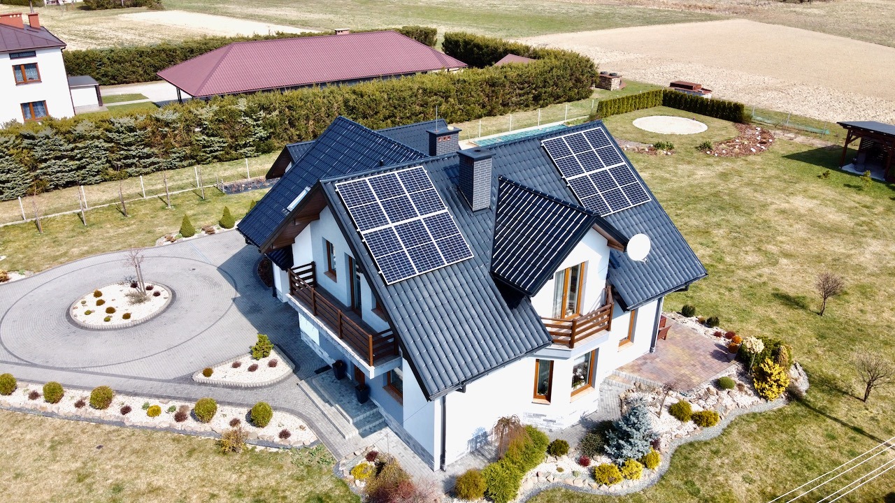 Widok z lotu ptaka na dom jednorodzinny z panelami słonecznymi na dachu, otoczony zadbanym ogrodem z okrągłym podjazdem z kostki brukowej i licznymi nasadzeniami.