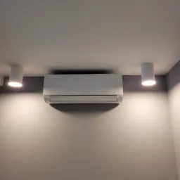 Biały klimatyzator Haier zamontowany na szarej ścianie, oświetlony dwoma cylindrycznymi lampami sufitowymi.