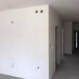 Wnętrze pomieszczenia po nałożeniu gładzi gipsowej. Widoczne otwory instalacyjne w ścianie i sufit, surowa podłoga, drzwi w tle.