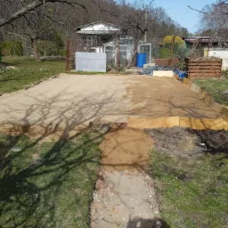 Przygotowany teren pod fundamenty z drewnianym szalunkiem, widoczne ślady cienia drzewa na piasku, w tle zabudowania gospodarcze i drzewa bez liści.