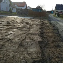 Przygotowany teren pod budowę z widocznymi śladami opon i wyrównaną powierzchnią ziemi, w tle zabudowa jednorodzinna i droga gruntowa.