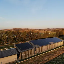 Widok z góry na budynek z panelami słonecznymi na dachu, otoczony zielenią i polami, z elementami infrastruktury w tle.