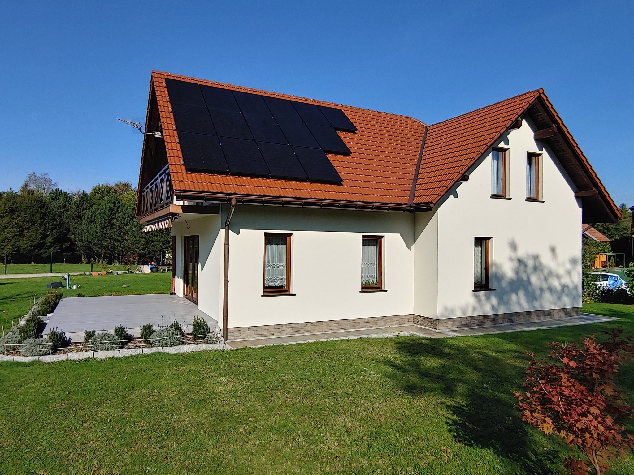 Dom jednorodzinny z brązowym dachem i panelami fotowoltaicznymi na połaci dachu, widoczne okna z firankami, taras z szarą deską kompozytową i ogród z trawnikiem.