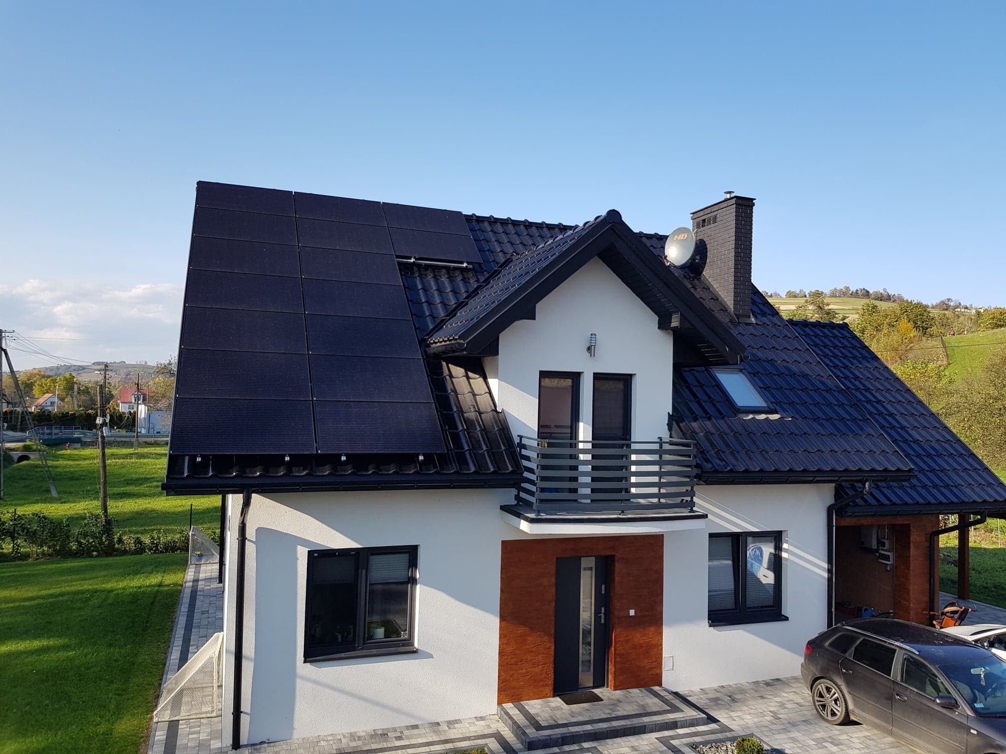 Nowoczesny dom jednorodzinny z panelami słonecznymi na dachu, widok z góry w słoneczny dzień, z samochodem zaparkowanym obok.