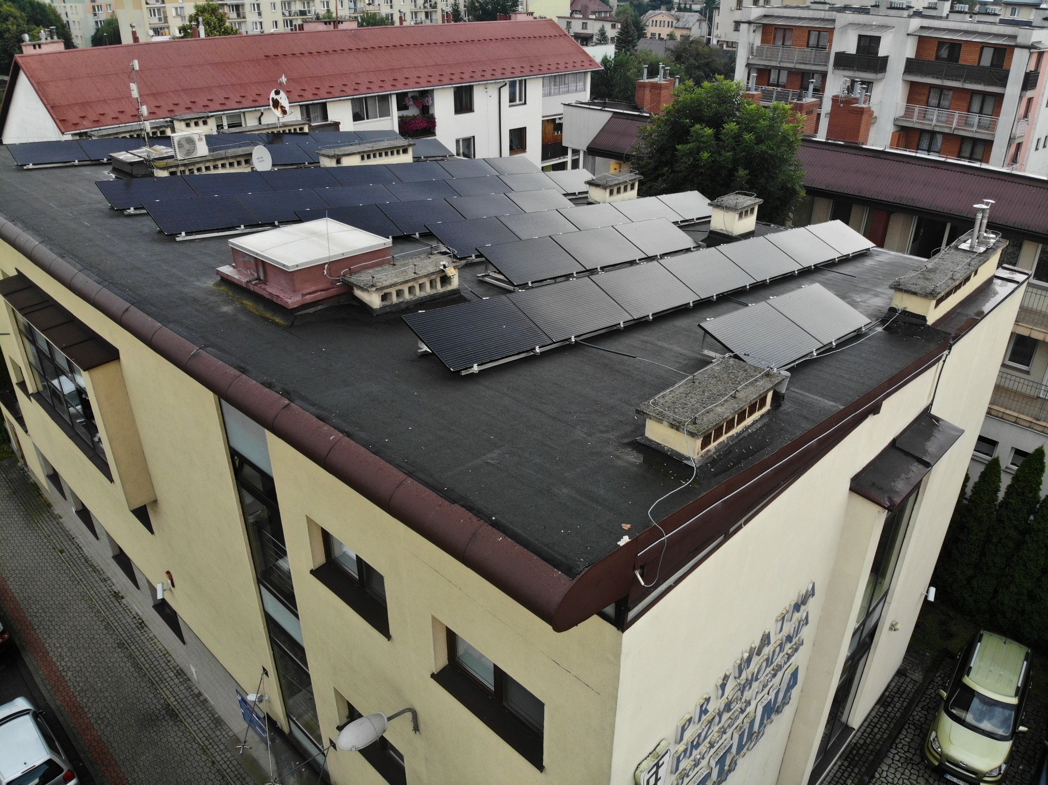 Widok z góry na budynek z panelami słonecznymi na płaskim dachu, widoczne również elementy wentylacyjne i kominy, w tle zabudowa miejska.