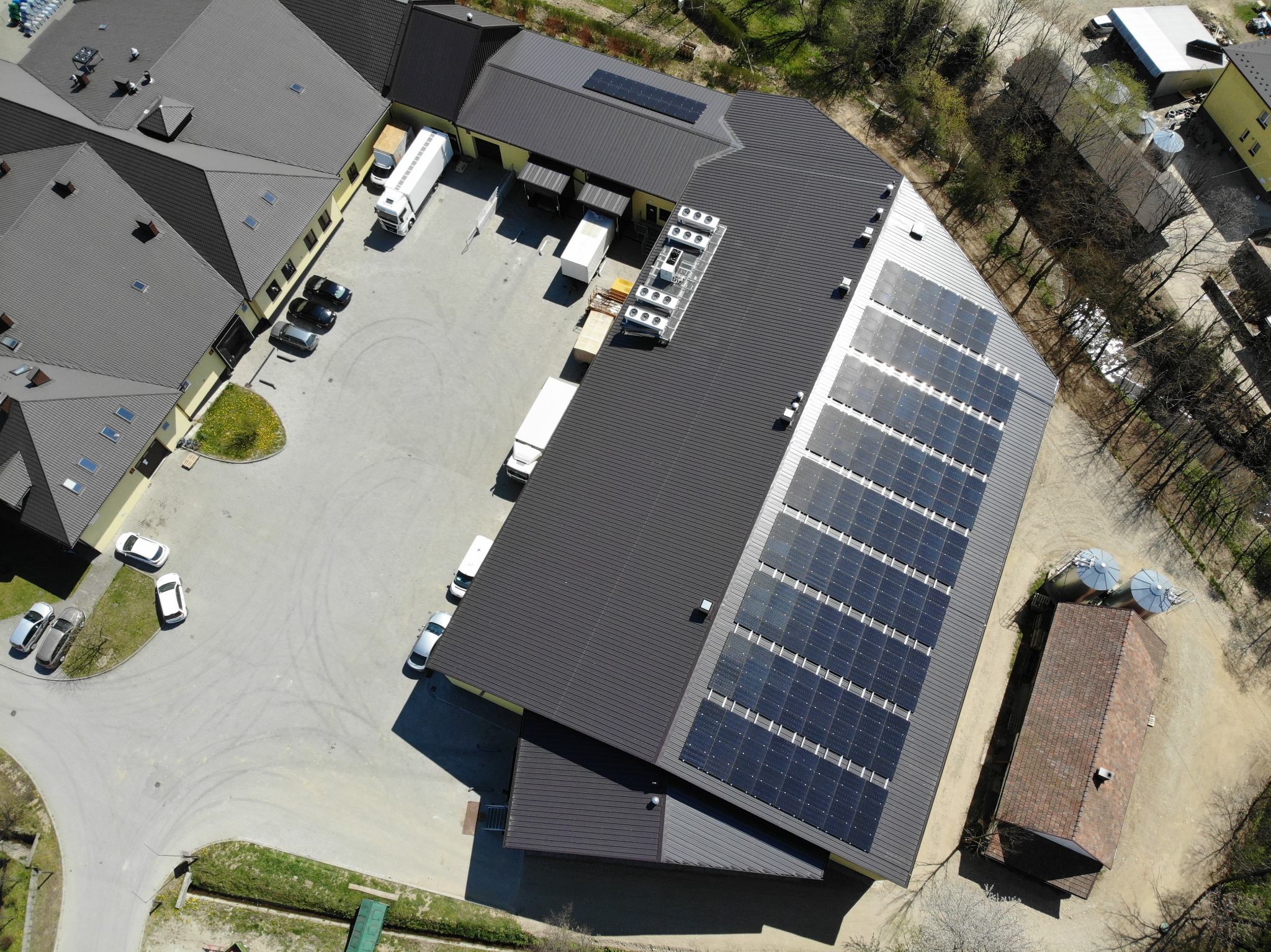 Widok z lotu ptaka na duży budynek z ciemnoszarą blachodachówką, na której zamontowano panele słoneczne. Obok budynku zaparkowane samochody i stoją samochody dostawcze. Widoczne także silosy...