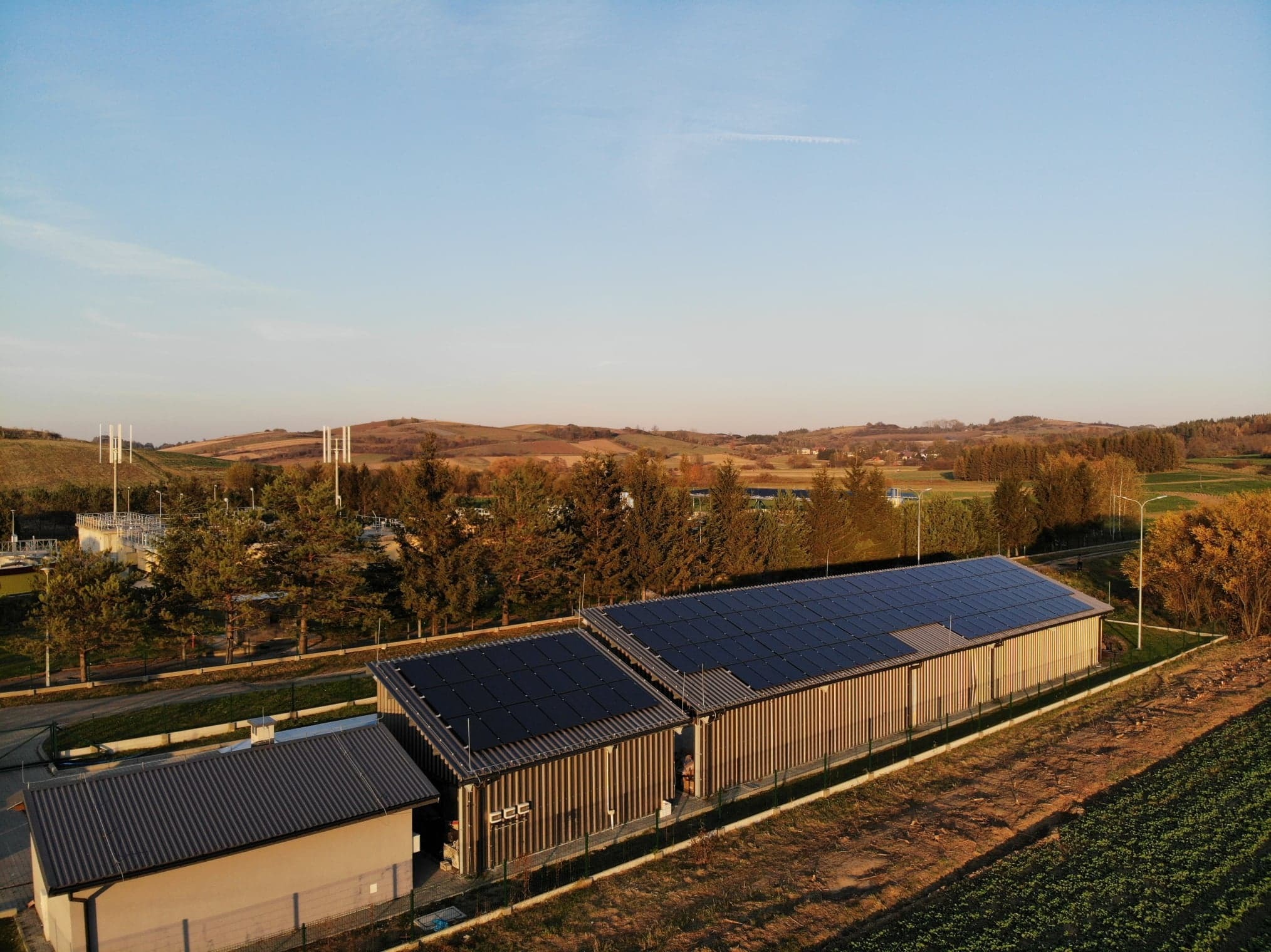 Widok z góry na budynek z panelami słonecznymi na dachu, otoczony zielenią i polami, z elementami infrastruktury w tle.