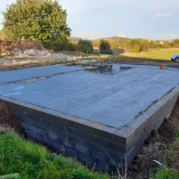 Wylany fundament z bloczków betonowych na działce, widoczne zbrojenie, w tle drzewa i zaparkowany samochód.