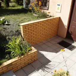 Ułożona jasna kostka brukowa na tarasie z niską ścianką z cegły, obok wejścia do budynku i rabaty kwiatowej.