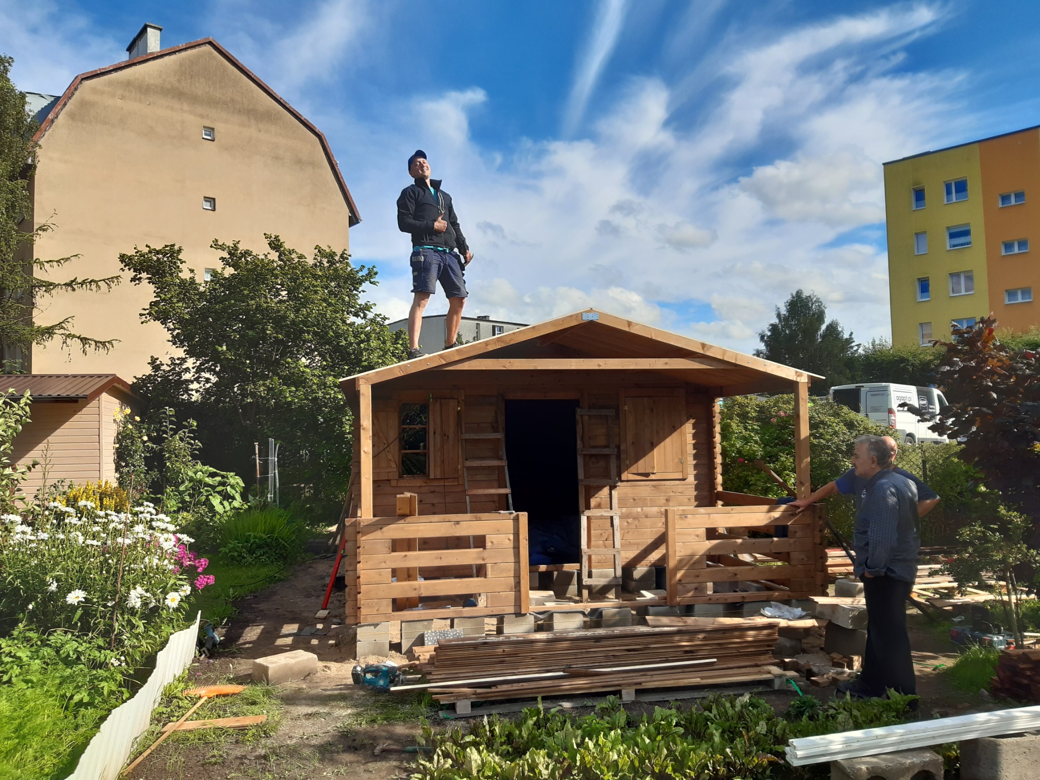 Mężczyzna w czarnej kurtce i niebieskich spodenkach stoi na dachu drewnianej altany w trakcie budowy, drugi mężczyzna obserwuje z dołu, w tle budynki mieszkalne i błękitne niebo z chmurami.