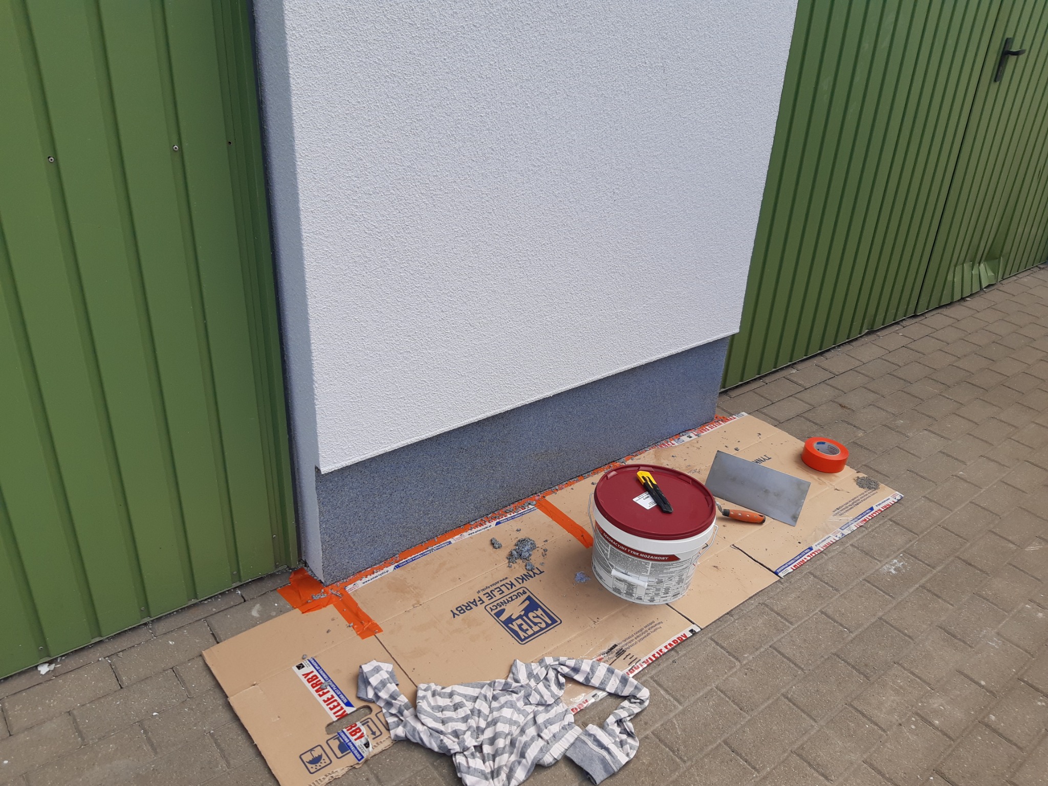 Przygotowania do nałożenia tynku mozaikowego na cokół budynku, widoczny kubeł z tynkiem, szpachelka, nóż do tapet i taśma malarska, zabezpieczone kartonem podłoże.