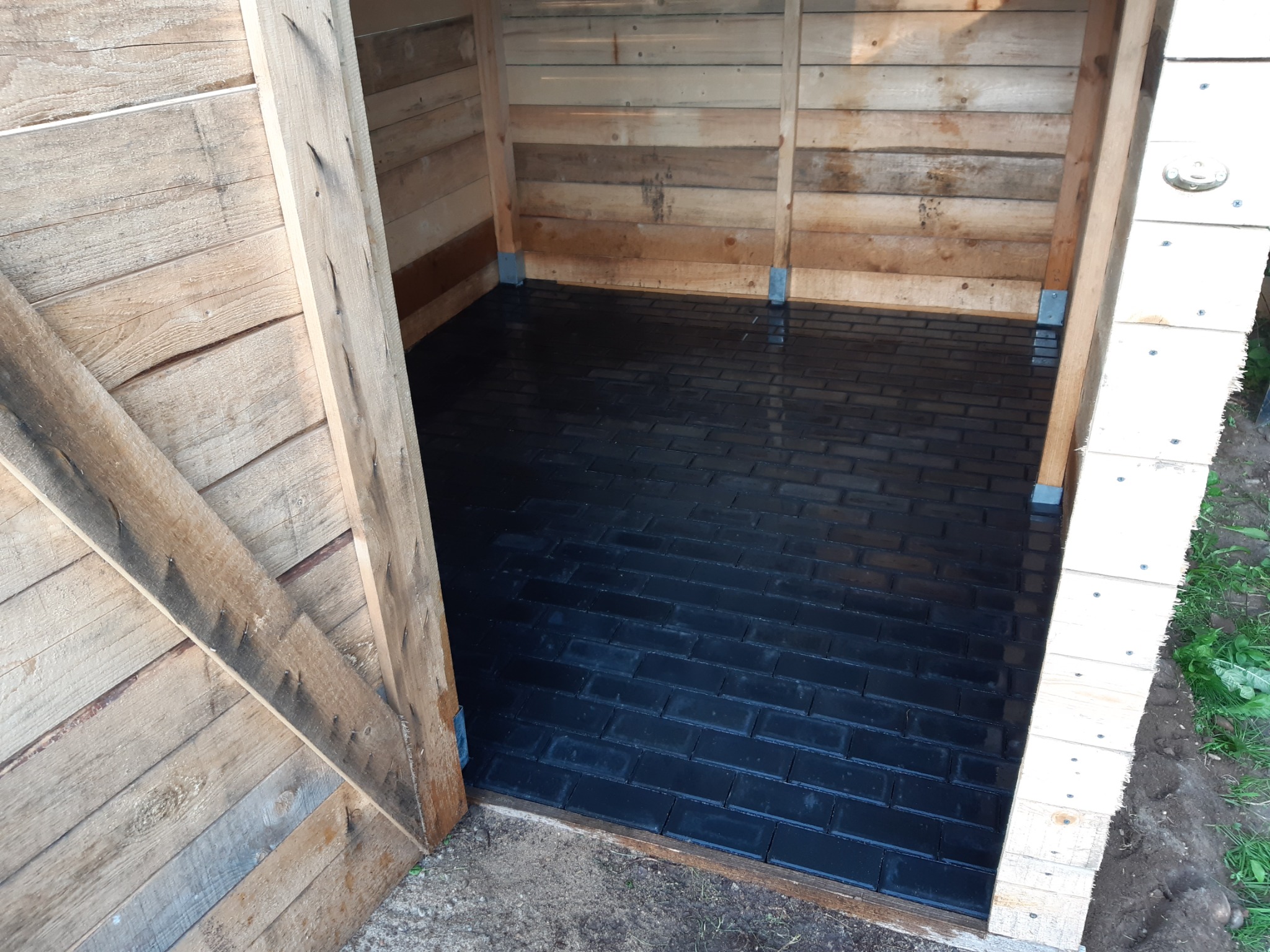 Wnętrze drewnianej szopy z nowo ułożoną, błyszczącą, czarną kostką brukową w jodełkę, widoczne niedoskonałości w ułożeniu przy ścianach.