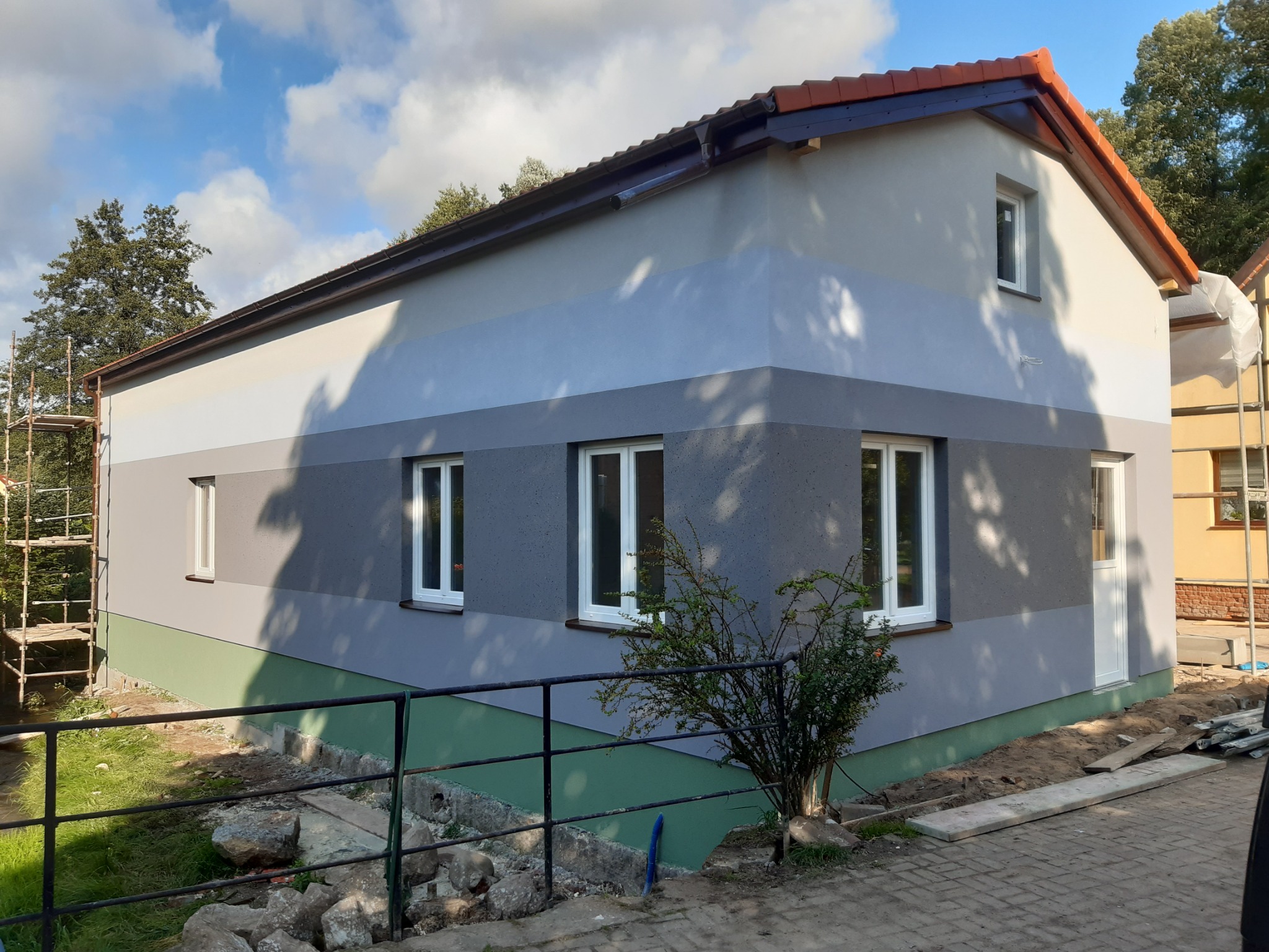 Świeżo otynkowany budynek z poziomymi pasami w odcieniach szarości, z widocznymi rusztowaniami i elementami przygotowania terenu wokół.