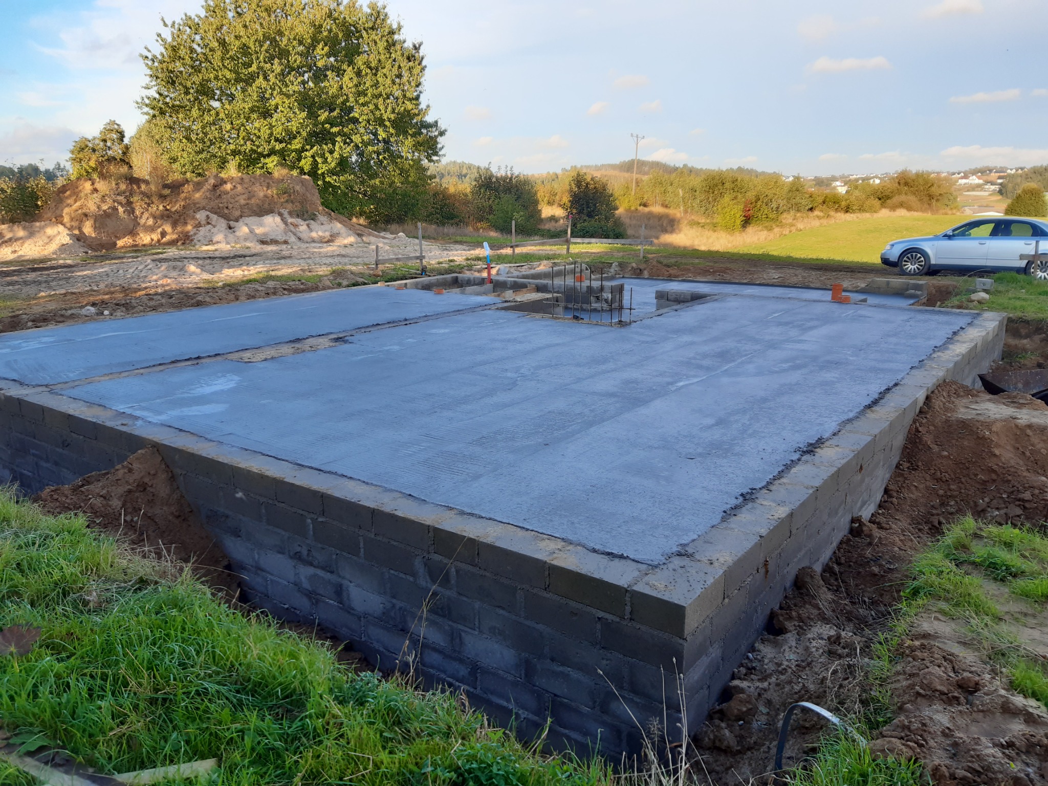 Wylany fundament z bloczków betonowych na działce, widoczne zbrojenie, w tle drzewa i zaparkowany samochód.