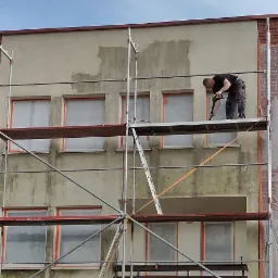 Pracownik na rusztowaniu czyści elewację myjką ciśnieniową, przygotowując ją do dalszych prac wykończeniowych. Okna zabezpieczone folią, widoczne fragmenty ceglanej elewacji i drewnianego ogrodzenia.