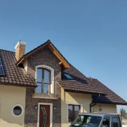 Nowoczesny dom jednorodzinny z elewacją w odcieniach beżu i brązu, z elementami cegły klinkierowej i brązową dachówką, na tle błękitnego nieba, z zaparkowanym szarym samochodem dostawczym firmy...