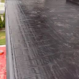 Czarna, pomarszczona membrana dachowa, prawdopodobnie EPDM, ułożona na płaskim dachu z widocznym połączeniem z obróbką blacharską i fragmentem elewacji w tle.