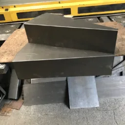 Dwa metalowe elementy o trójkątnym przekroju, ułożone jeden na drugim na warsztatowym stole, w tle widoczna maszyna do obróbki metali.