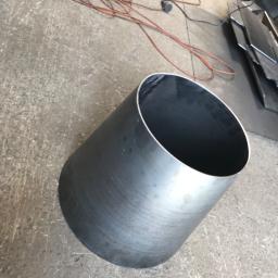 Auto-remont Łukasz Wójcik - Cylindryczny element z surowej blachy stalowej, widoczny na tle betonowej posadzki warsztatu, z fragmentami kabla zasilającego i arkuszami blachy w tle.