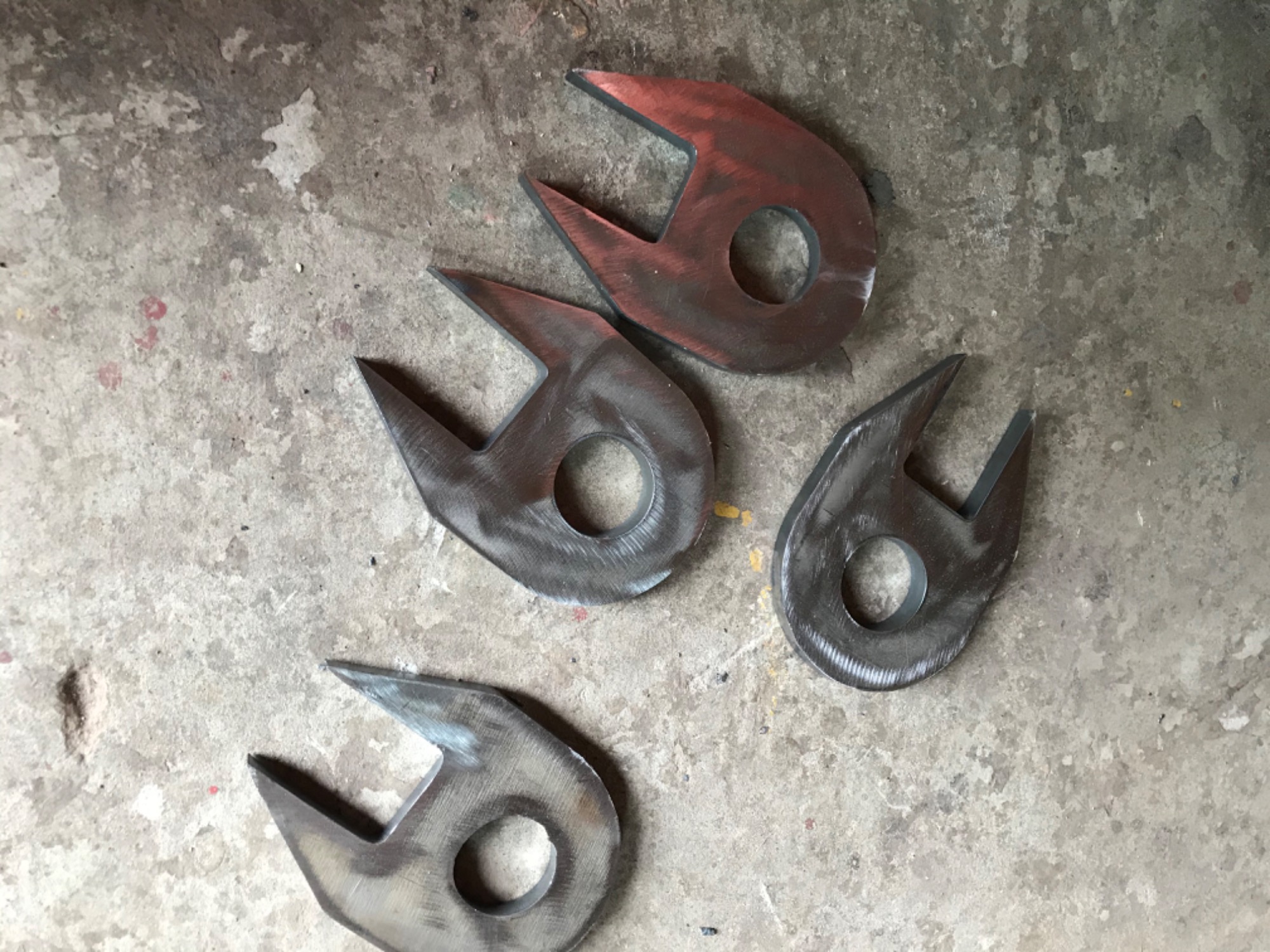 Cztery metalowe elementy z otworem i dwoma ostrzami, leżące na betonowej posadzce, widoczne ślady obróbki.